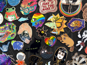 Custom enamel pins by Alchemy Merch featuring colorful artistic designs including animals, butterflies, skulls, and pride pins